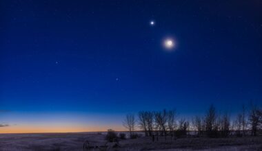 The moon shines in the evening sky with a bright star-like planet glowing close to its upper left above a snowy field lined with trees. The glow of the setting sun can be seen brightening the horizon.