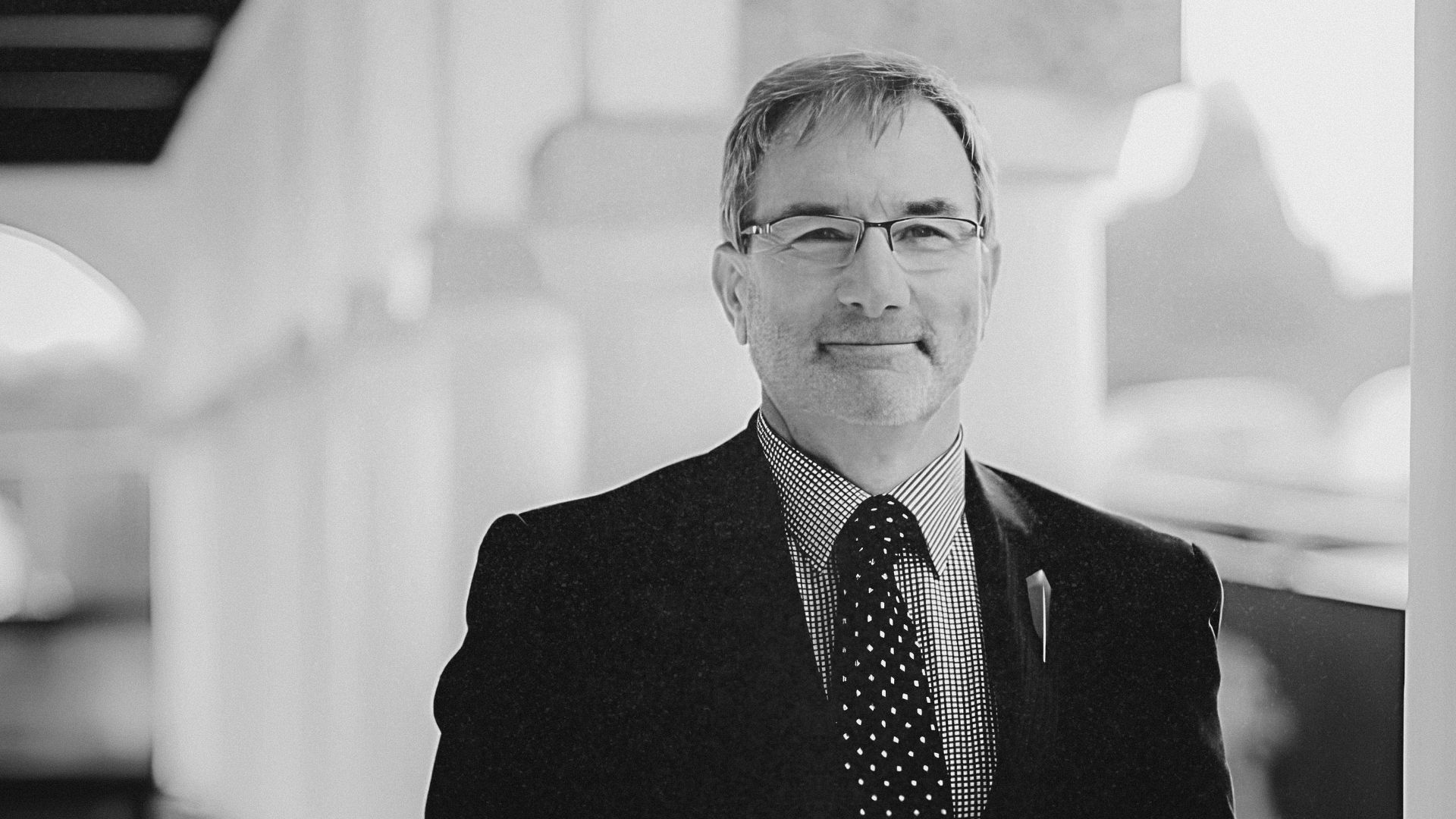 A man in a black suit, white shirt, and black polka-dot tie stands in a hallway, wearing glasses and a pin on his lapel.