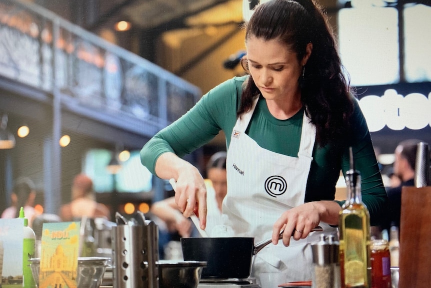 Hannah wears an apron, long sleeve and has hair in pony tail. She whisks in a bowl in a kitchen.