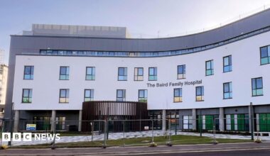The hospital is a large, four storey white and grey building, part of the building is blue and extends from the front of the hospital.