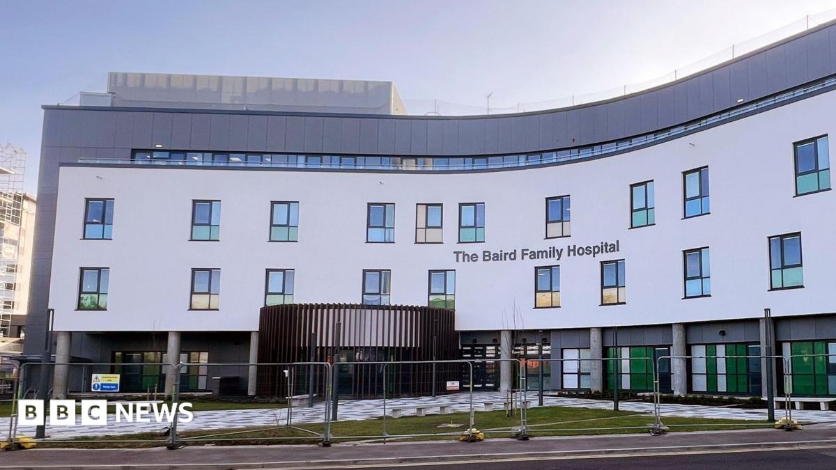 The hospital is a large, four storey white and grey building, part of the building is blue and extends from the front of the hospital.