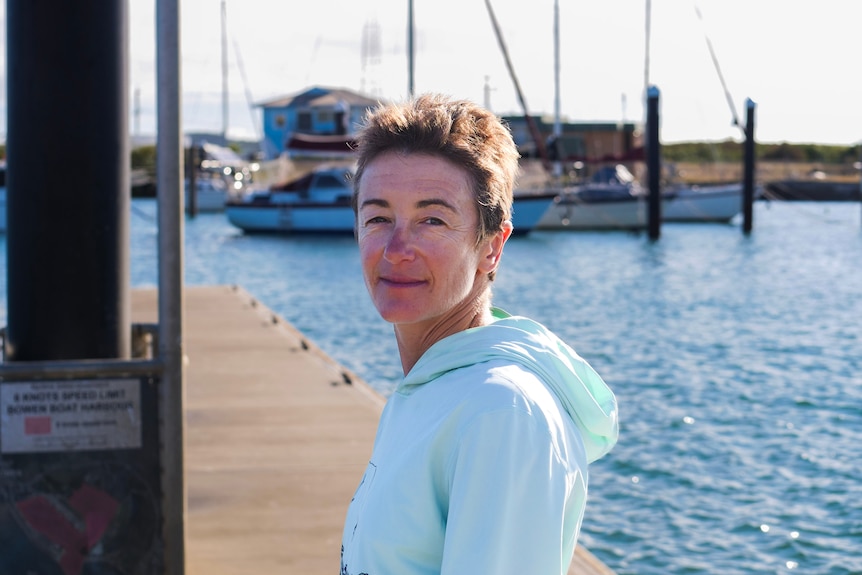 A woman with short, golden-brown hair wears a light hoodie as she stands on a pier at a marina.