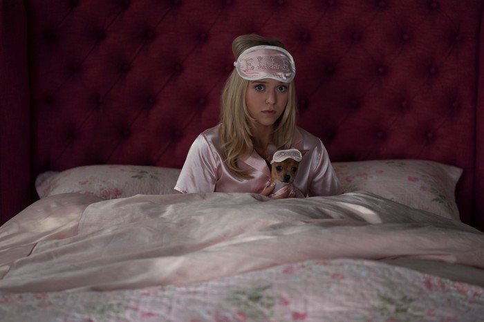 Elle - First Look Lexi Minetree as Elle Woods in Elle sitting in bed, wearing pink satin pyjamas and an eye mask with her chihuahua in her lap in a matching Elle mask.