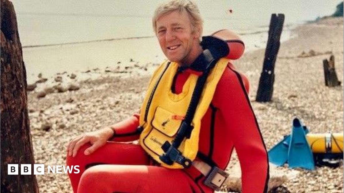 A man with fair hair sitting on a pebble beach. He has a red wetsuit on and a yellow life vest over the top. He is smiling as he looks into the camera, sideways facing.