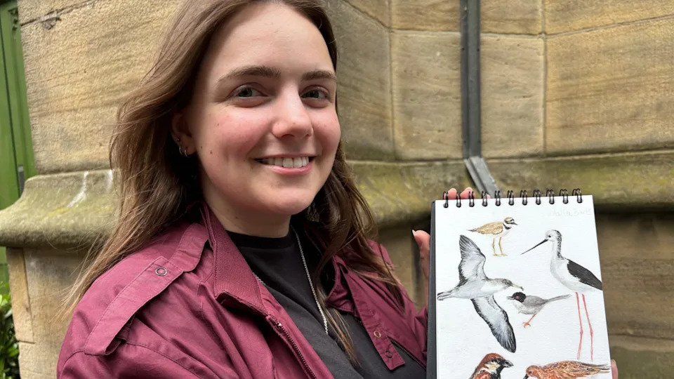 A smiley woman with dark hair and a purple jacket holds up a book of hand drawn pictures of birds.