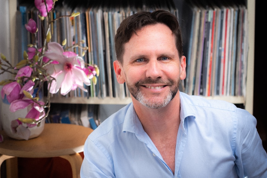 Robbie Buck sitting in front of a crowded record shelf, with a table with orchids in a vase on it to the left of frame.