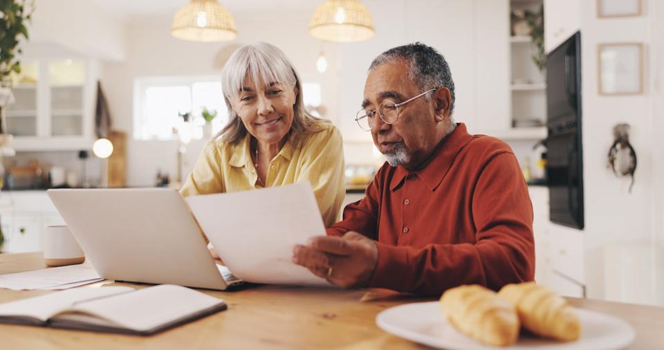 A multiracial senior couple sits at a wooden kitchen table, reviewing paperwork and looking at a white laptop. The woman, with grey hair and a yellow shirt, smiles slightly while looking at the man. The man, with grey hair, a beard, glasses, and an orange polo shirt, holds up a white document and points to it while looking at it intently. A white coffee mug, an open notebook, and two croissants on a white plate are also visible on the table. The background shows a modern, bright kitchen with hanging light fixtures.