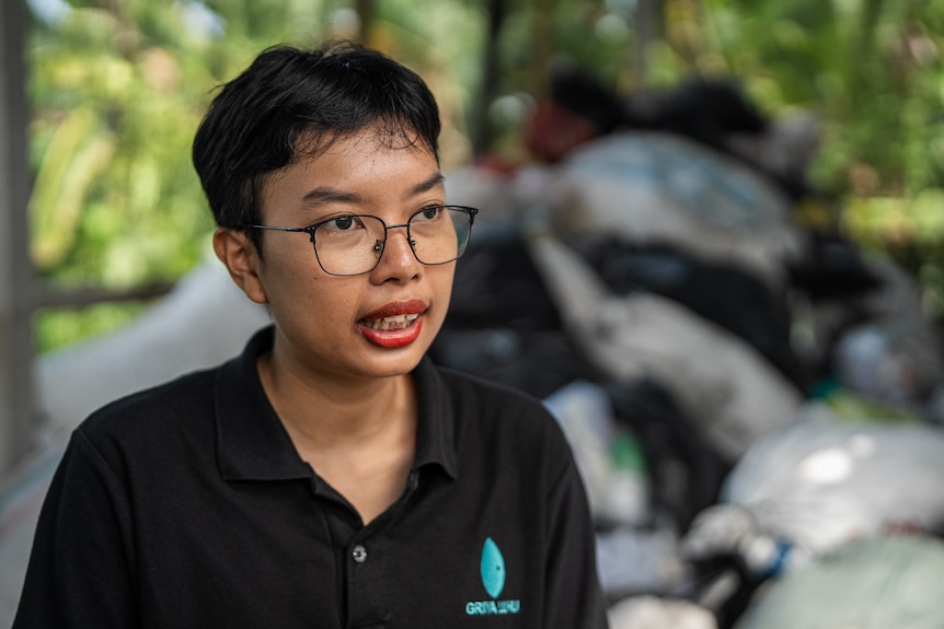 An Indonesian wearing glasses and a black polo top sits near some trash.