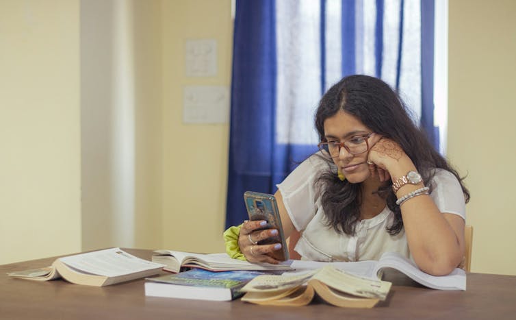 A woman with long hair and glasses stares at her phone surrounded by books on a desk for studying