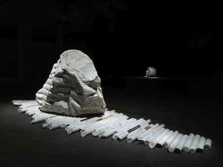 Large glacial erratic boulder displayed on drill cores in a dark gallery. Metal rods placed among the cores reference metal resources extracted from the Earth.