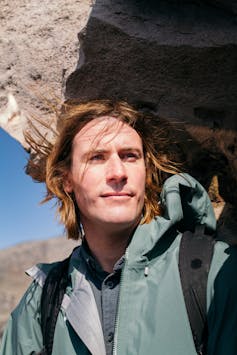 Portrait of artist Julian Charrière standing beside a rocky outcrop in a mountainous landscape. He wears a green jacket and backpack and looks into the distance.