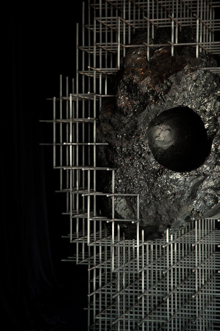 Large black lump of coal suspended within a steel scaffold structure in a dark gallery space. A smooth, head-sized cavity is carved into the coal, inviting visitors to place their head inside the sculpture.