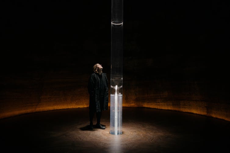 Artist Julian Charrière stands beside a tall transparent column inside a circular, dimly lit chamber. The illuminated installation, Breathe, extends from floor to ceiling and is designed to release oxygen extracted from ancient rocks.