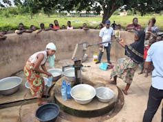 A group of people stands around a borehole collecting water in basins