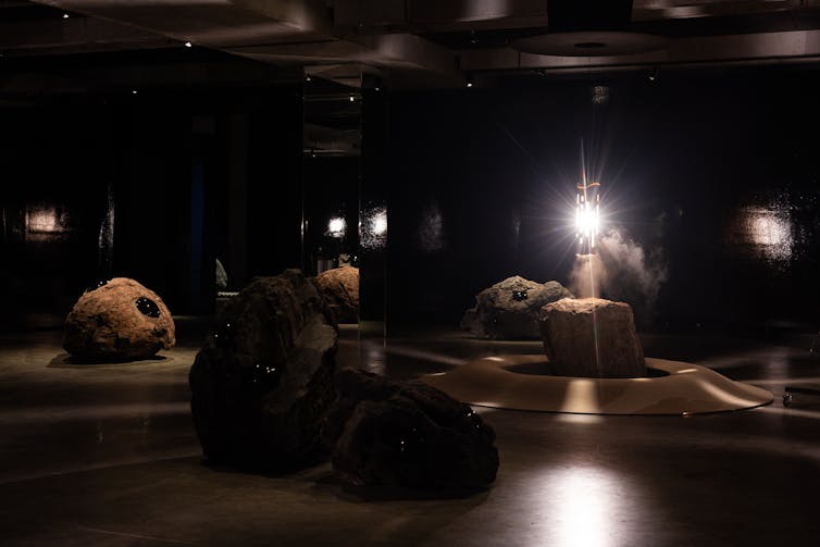 Installation view of Hard Core by Julian Charrière at MONA. Large volcanic rocks and lava bombs are arranged in a dark, mirrored gallery around a bright central light source, creating the impression of a volcanic chamber.