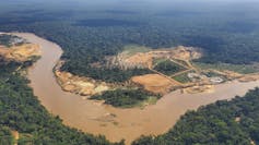 An aerial photo showing a very brown river and a large patch of green forest eaten away by a mine