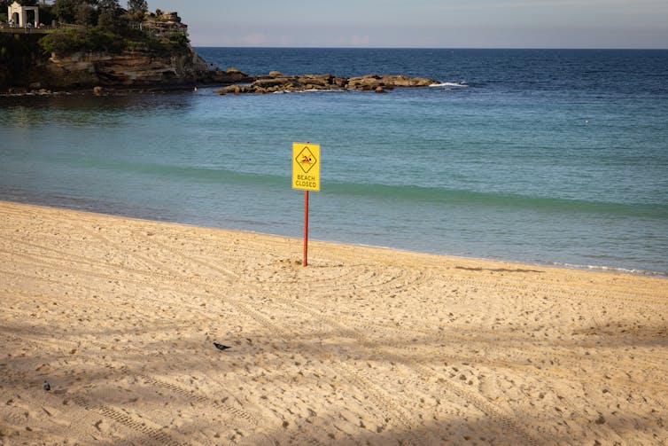 sign saying beach closed, empty beach and calm sea behind.