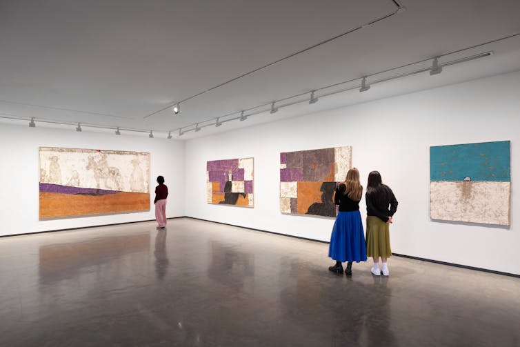 A large white gallery. Three people look at pastel paintings.