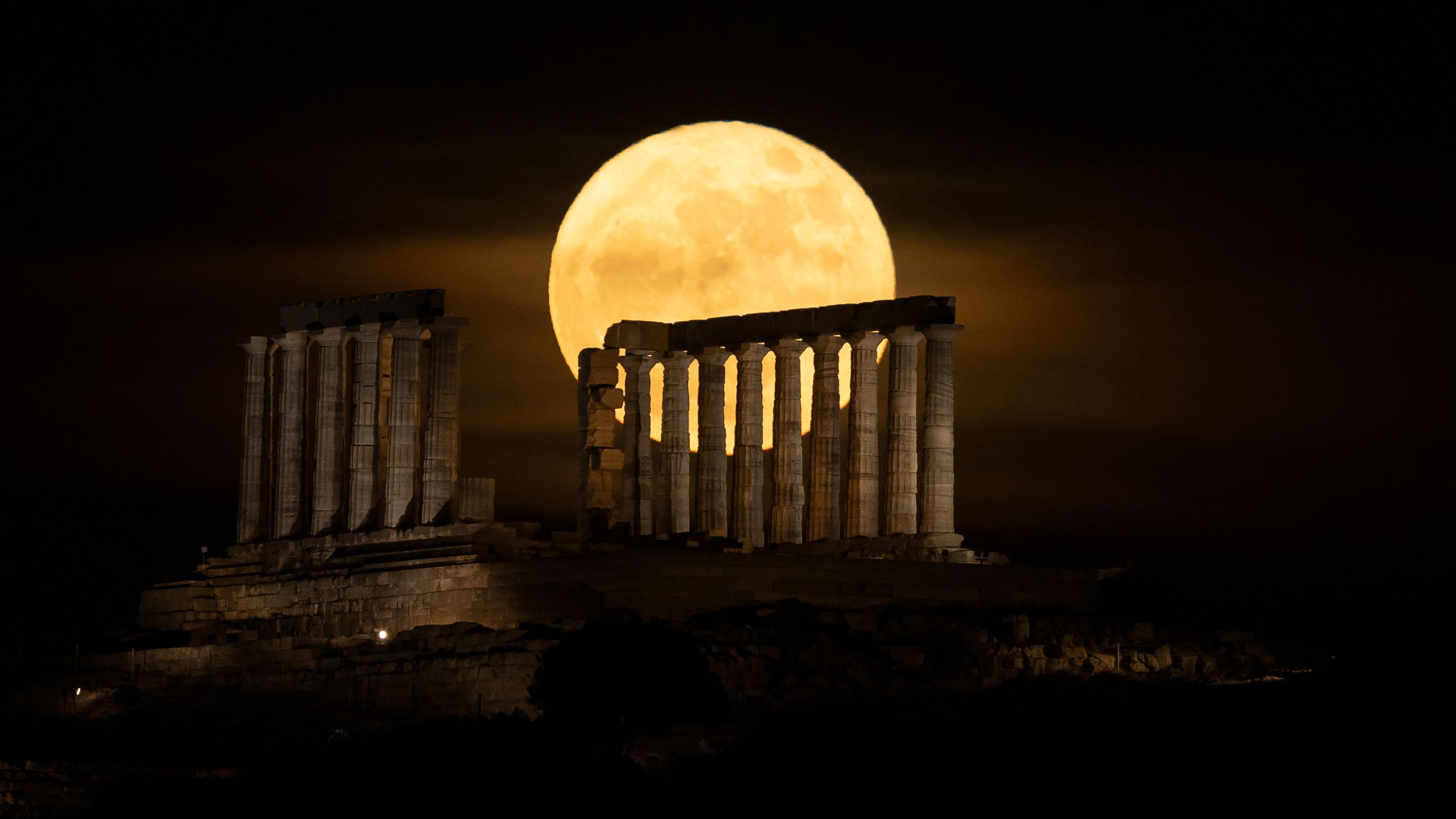 A yellow full moon glows in a dark sky behind the columns of an ancient ruined temple.