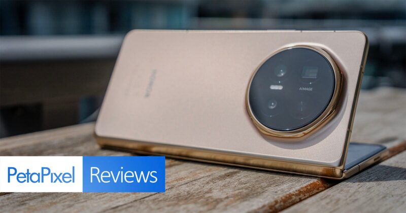 A gold-colored smartphone with a large circular camera module is propped up on a wooden surface. The “PetaPixel Reviews” logo appears in the bottom left corner of the image.