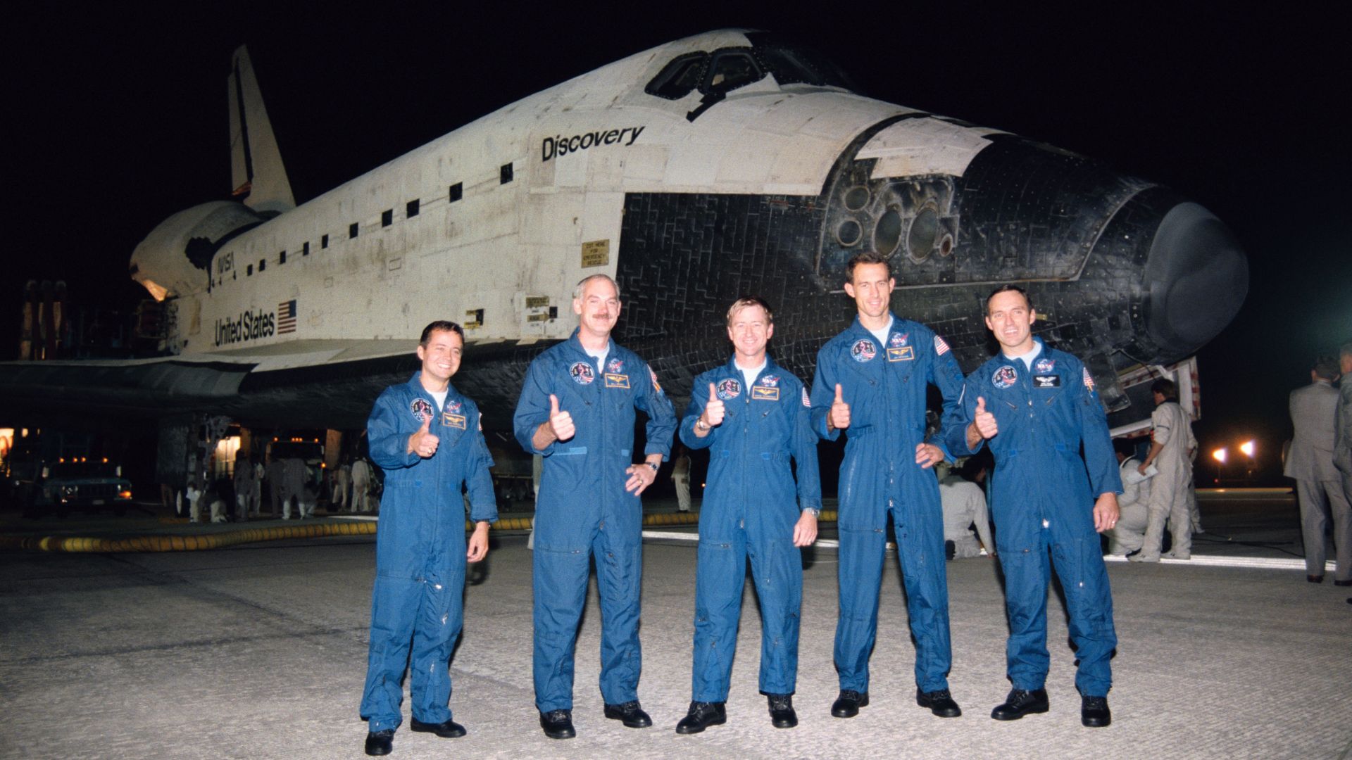 A crew of five astronauts in blue flightsuits stand giving the thumbs up in front of a space shuttle at night