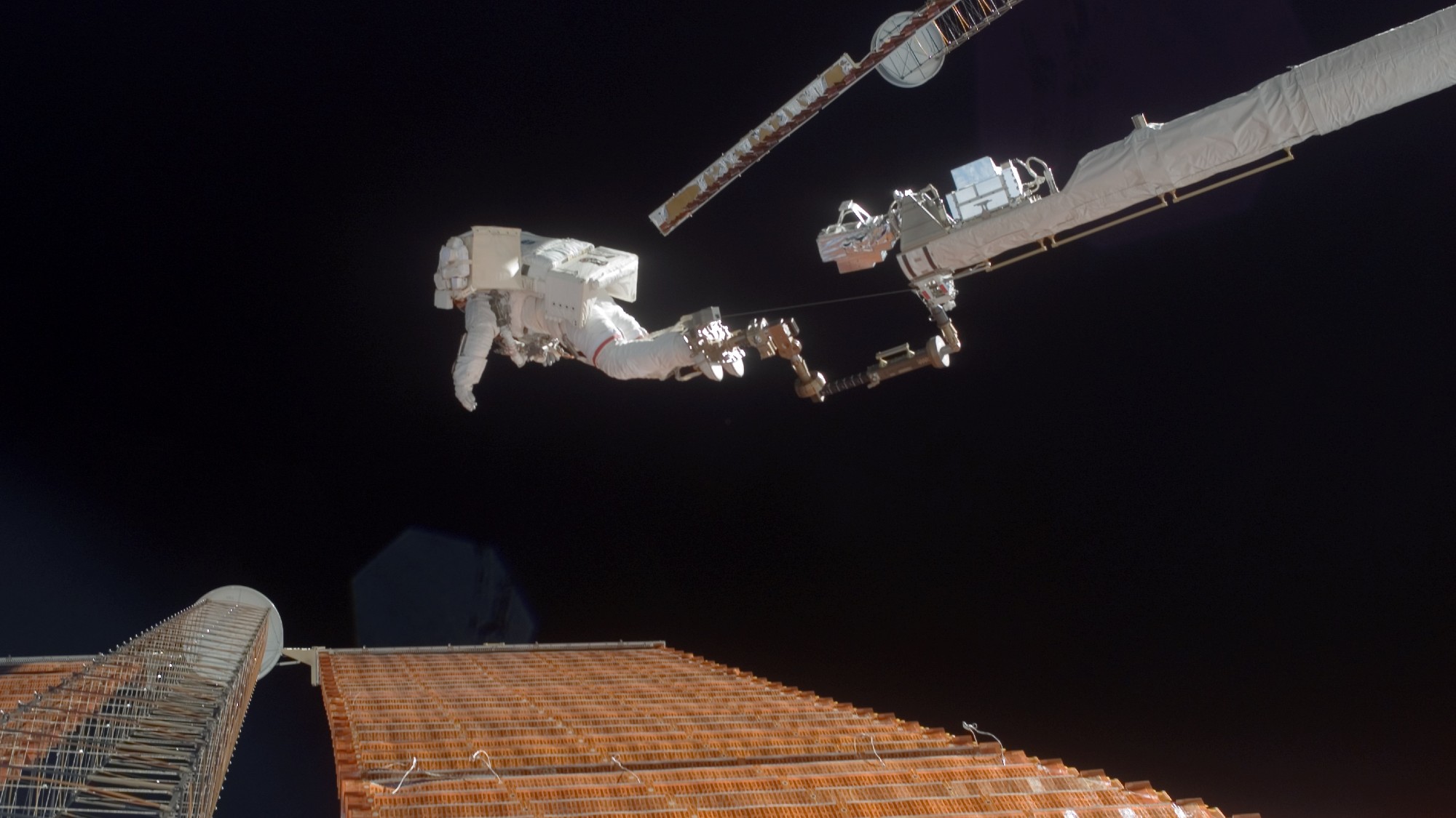 an astronaut in a bulky white spacesuit on the end of a robotic arm above large rectangular solar panels