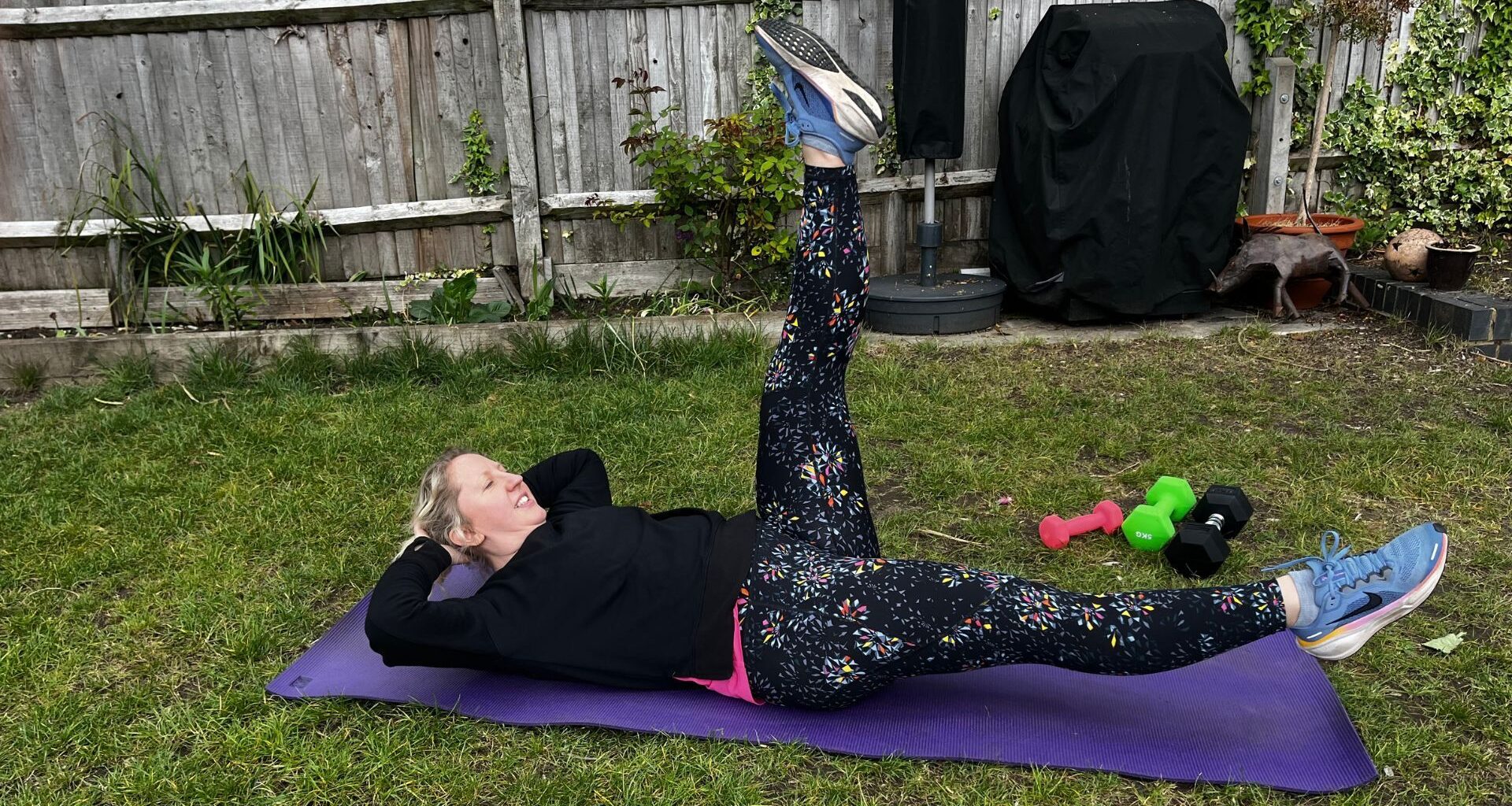 Kat Storr doing Pilates scissors exercise on yoga mat in garden