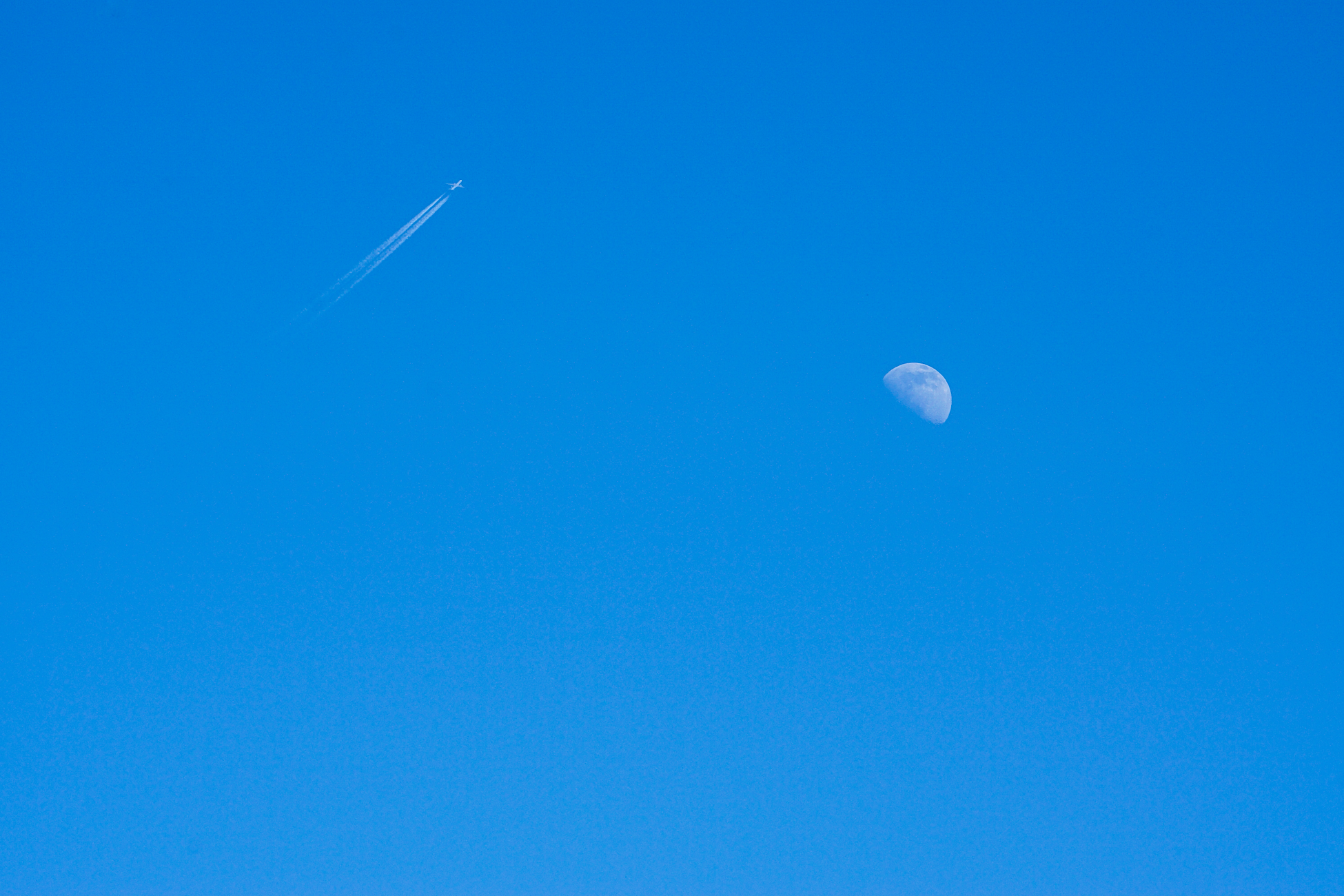 A high-altitude jet leaves a contrail near a visible moon in the clear blue sky.