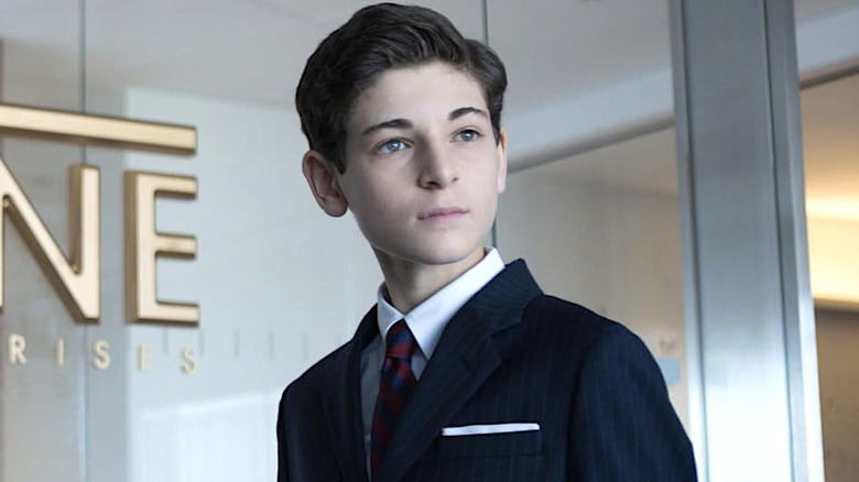 Bruce Wayne in a suit standing in front of a Wayne Enterprises glass window in 
