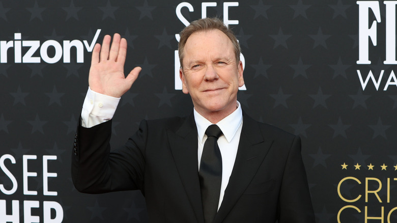 Actor Kiefer Sutherland waving to the camera in front of a black screen with gray stars and sponsor names on it
