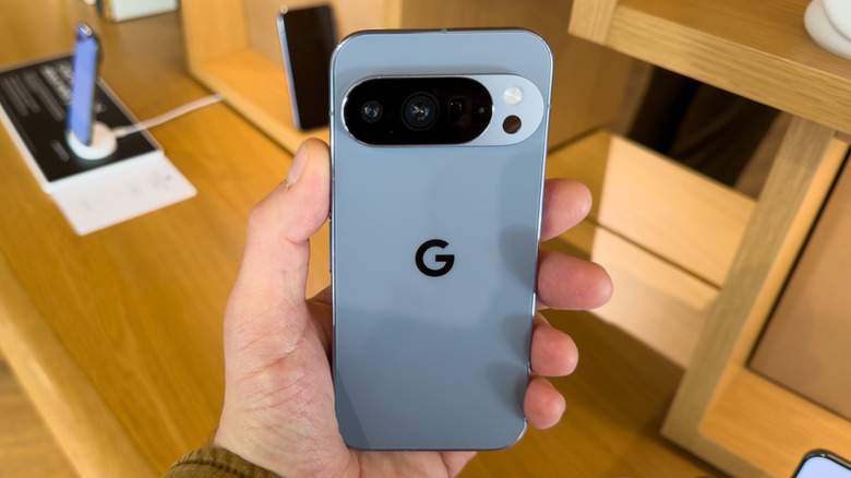 A Google Pixel 10 sitting in a user's left hand, the device appears to be attached to a security device inside a store