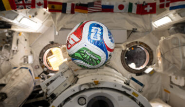 A white, red, blue, and green soccer ball floats inside the International Space Center. The FIFA logo is visible in the blue part of the ball facing the camera. The area in the background is mostly white, with a banner of country flags at the top of the photo.