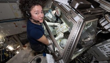 Jessica Meir wears a navy-blue T-shirt and a headset. She looks at the camera while her arms are inserted into the Life Sciences Glovebox. She holds a rectangular plate with multiple sealed syringes and tubes. There are many bags within the glovebox with lab supplies. Metallic surfaces and electronics surround the area outside the glovebox.