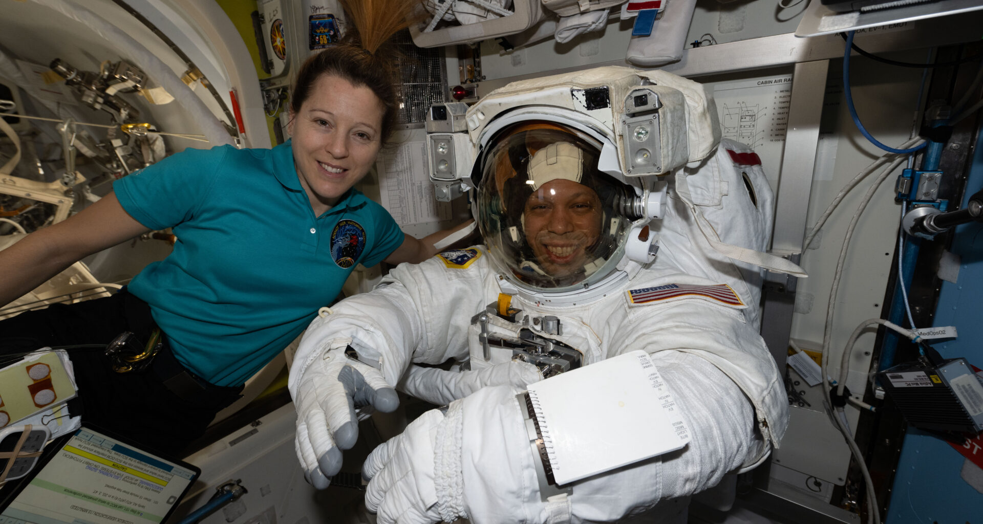 ESA astronaut Sophie Adenot and NASA astronaut Chris Williams smile at the camera. Adenot wears a teal polo shirt with a patch on the front. Williams is in a white spacesuit. They are on the International Space Station.