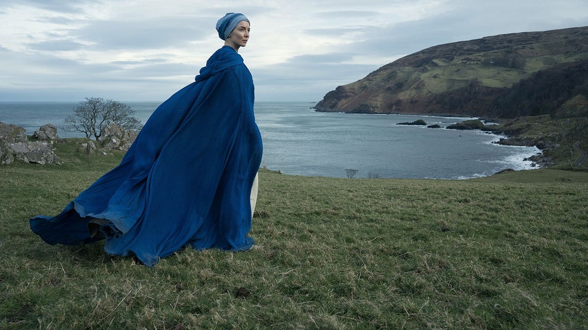 Jodie Comer wearing a blue cloak standing on a grassy coastal hillside overlooking a bay.