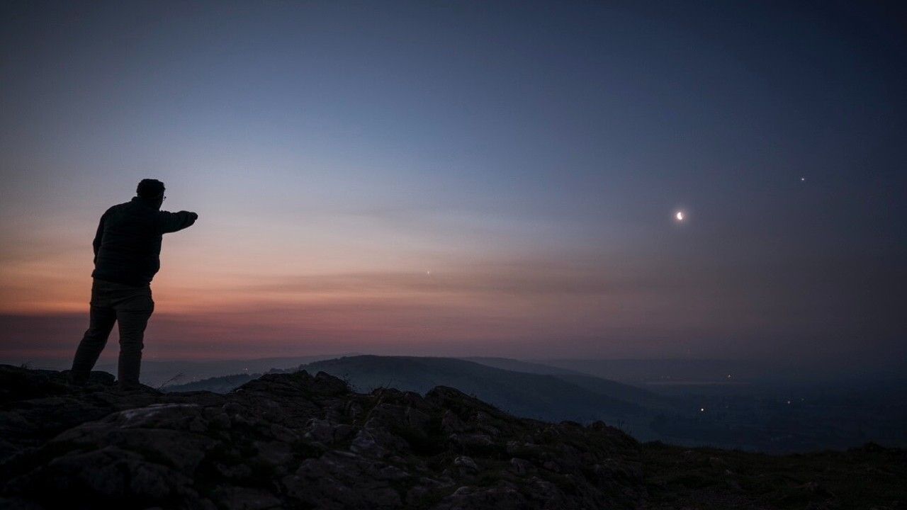 a person stands with their back to the viewer, pointing at several bright dots in the early morning sky