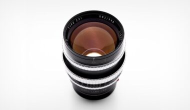 A camera lens with a black body and silver focus rings is shown from above on a white background. The glass elements reflect light, and text around the lens edge reads “TYPE Z1 LIGHT LENS LAB.”.