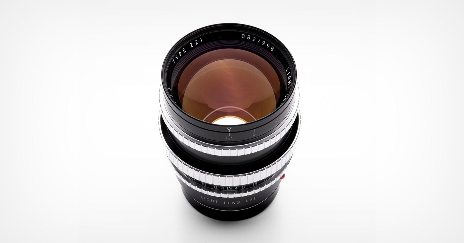 A camera lens with a black body and silver focus rings is shown from above on a white background. The glass elements reflect light, and text around the lens edge reads “TYPE Z1 LIGHT LENS LAB.”.