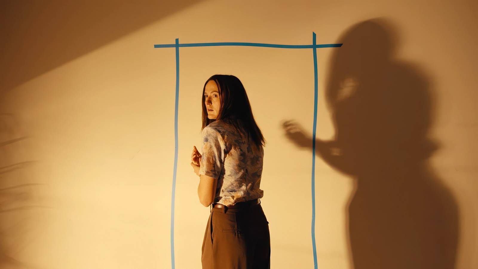 Mary (Renate Reinsve), a thin, pale woman with long black hair, stands with her back to the camera, looking nervously back over her shoulder, in front of a tape outline of a doorway on a blank wall in Backrooms