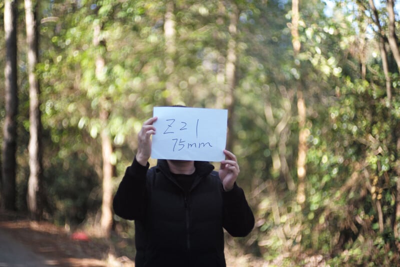 A person outdoors holds a white paper in front of their face with "ƒ2.2 75mm" handwritten on it. The background is a sunlit, blurry forest path with green foliage.