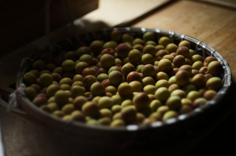 A round woven basket filled with small, yellow-green and reddish fruits sits on a wooden surface in soft, diffused light.