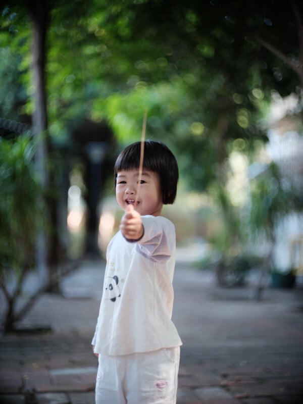 A young child with short black hair, wearing a white shirt and shorts, smiles while holding a stick towards the camera in an outdoor, tree-lined setting.