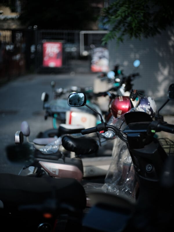 A row of parked scooters and motorcycles on a shaded sidewalk, with the focus on a rearview mirror and the background softly blurred.