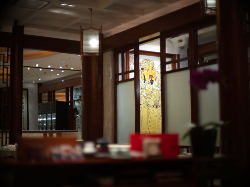 A warmly lit interior of an Asian restaurant features wooden dividers, frosted glass panels, and a colorful traditional painting of a figure holding a fish on the wall. A blurred table with items is visible in the foreground.