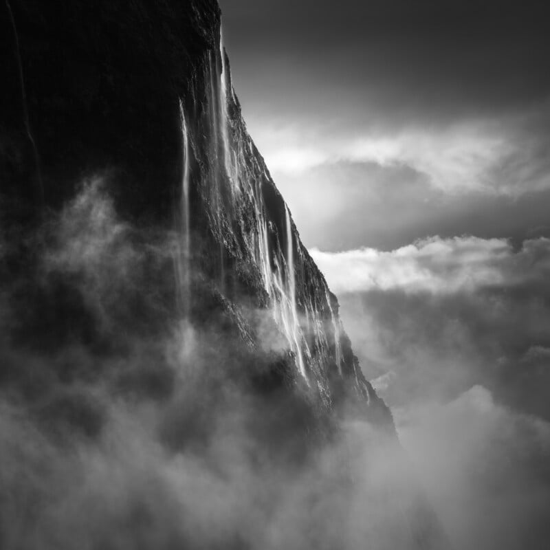 A dramatic black-and-white image of a steep, rocky mountain face with thin waterfalls cascading down, shrouded in mist and clouds, creating a mysterious and ethereal atmosphere.