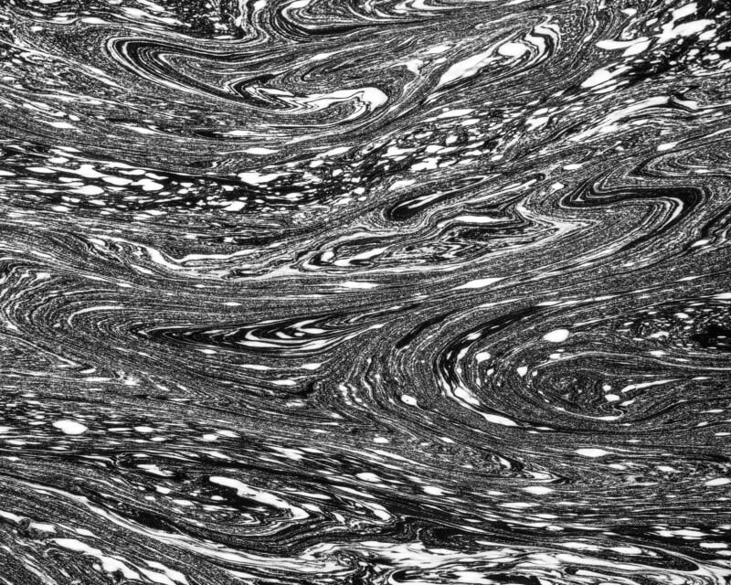 Black and white abstract image featuring swirling, marbled patterns with flowing lines and scattered white spots, resembling liquid or ink in motion.