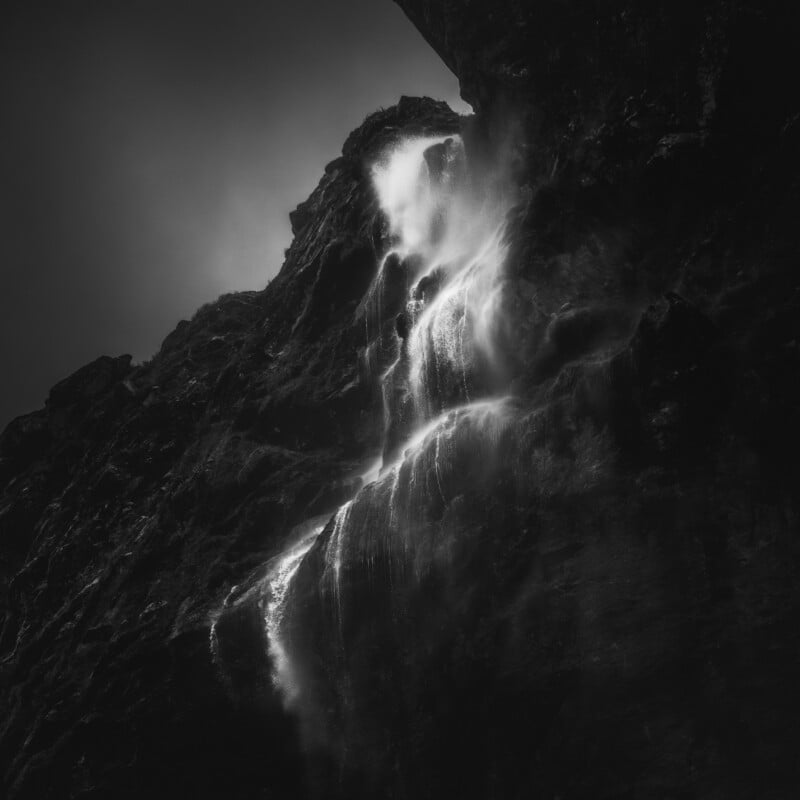 A dramatic black and white photo of a waterfall cascading down a rugged, dark rock face, with mist rising from the falling water and contrasting shadows creating a moody, mysterious atmosphere.