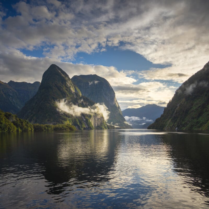 A serene lake reflects green, rocky mountains and a cloudy blue sky. Mist drifts in front of the mountains, and sunlight breaks through the clouds, creating a peaceful and dramatic landscape.