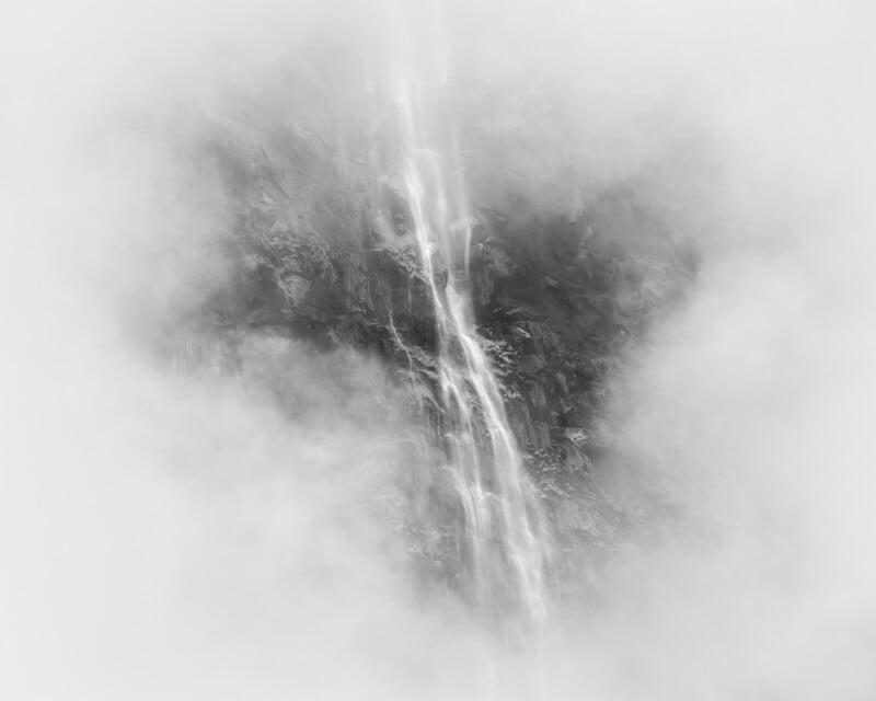 A tall, narrow waterfall cascades down a rocky cliff, partially obscured by thick mist or fog, creating a soft, ethereal, black-and-white scene.