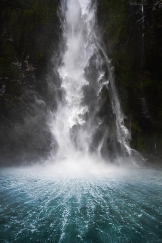A powerful waterfall cascades down a rocky cliff into a vibrant turquoise pool, creating mist and ripples on the water’s surface.
